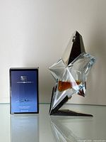 View of 50ml Angel Eau de Parfum bottle mounted on a star-shaped silver metallic stand next to the boxed 5ml miniature bottle on a reflective surface.