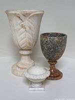 All three carved stone pieces grouped together: large alabaster urn with leaf motifs, multicolor marble goblet, and small white urn with bird figure