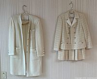 Two cream tweed suits hanging side by side: long coat with matching skirt on left, double-breasted jacket with pleated skirt on right.