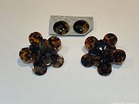 Two large tortoiseshell floral earrings and a pair of round stud earrings on a neutral background with hand for scale.