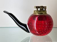 Side view showing the red art glass spherical body with embedded bubbles and black glass pipe stem extending to the left, topped with brass lighter mechanism.