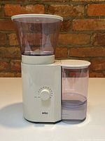 Photo showing the white Braun KMM 30 coffee grinder with two transparent compartments and timer dial on front, captured against brick background.
