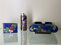 Photo of the set showing matchbox holder, lighter sleeve, and ashtray with clear cloisonné floral decorations on blue enamel backgrounds, showing some enamel chips and slight bend on matchbox holder.