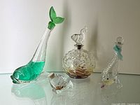 Four vintage fish shaped perfume bottles displayed on a reflective surface against a white background: a tall bottle with green tail stopper and turquoise liquid, a round bottle with bubble texture and amber liquid, a long slender bottle with ribbon tied near neck, and a small bottle with beige cap.