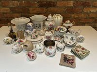 Full view of the lot showing various porcelain and ceramic potpourri balls, dishes, and decorative containers arranged on a white table against a brick wall.