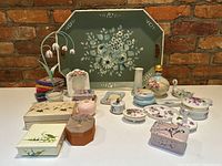 Wide image showing all items in the lot including decorated tray, pretty ceramic and porcelain trinket boxes with floral patterns, photo frame, decorative books, and small vases and figurines arranged on a white table against a red brick wall.