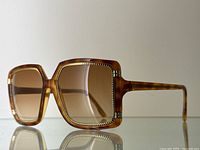 Three-quarter front view showing oversized square frames and gold-tone braided trim.