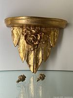 Front view of semi-circular gold leaf wooden wall shelf with carved rose and leaf pattern and two small metal floral hangers placed in front.