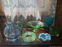 The wide view of the lot showing all seven glass items including the uranium glass pedestal bowl, blue pitcher, two green goblets, blue ruffled bowl, green square bowl, and green ring-shaped item on a wooden surface under window light.