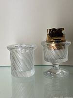Polished and satin twist crystal table lighter and cigarette jar by Lalique shown side by side on glass shelf with neutral background.