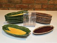 View of green Enesco corn on the cob plates stacked on left with a set of brown McCoy plates stacked on right, and set of four metal corn-shaped holders in center showing lot contents.