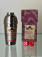 Miniature cocktail shaker with Old Quaker label next to original box and two dice on reflective surface.