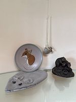 Full view of all four items on a shelf: plate, hanging glass mouse, resin sculpture, and grey jewelry pouch.