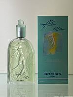 Photo shows clear rectangular glass perfume bottle with embossed female figure design next to its original box. Box has green-blue painted design with dark blue 'ROCHAS' text and 'fleur d'eau' cursive script.