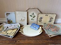 Overview of all trays and cutting boards laid out on wood floor against wallpaper background. Shows various designs including floral, botanical, bird, abstract, and village scenes.