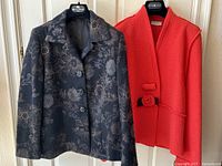 Two women’s jackets hung on hangers – orange felted wool jacket with flower closure and black wool jacket with floral pattern and leather buttons.