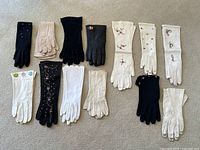 12 pairs of women's vintage gloves arranged in two rows on a carpet, showcasing various embroidery and colors including black, white, and beige.