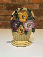 Full front view of the vintage wrought iron door stopper featuring a basket with colorful pansy flowers in a brick background.