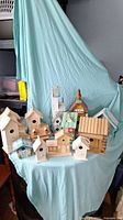 Photo of thirteen wooden bird houses of various shapes and sizes arranged on a blue draped surface. Includes log cabin style, pitched roofs, and natural wood finishes.