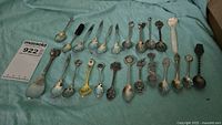 Full collection of 24 assorted souvenir spoons laid out on a blue fabric surface.