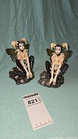 Two Harlequin fairy figurines seated on playing card themed bases with a MaxSold tag