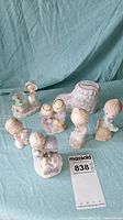Eight assorted Precious Moments porcelain figurines arranged on blue cloth, visible details show typical soft pastel colors and gentle expressions, include children and a flower-pattern shoe.