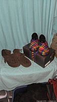 Pair of Manitoba Mukluks brown suede moccasin boots with fringe next to multi-colored Bernie Mev woven slip-on shoes on shoe box.