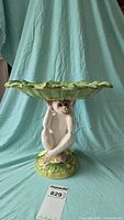 Front view showing white porcelain monkey holding a green leaf-shaped plate overhead, standing on a round base with foliage decoration.