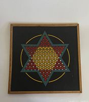Front view of wooden square Chinese Checkers board with star pattern and colored sections yellow, red, blue on black background with wooden frame.