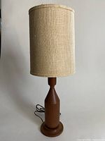 Image of the full lamp showing the wooden base with turned geometric design and the cylindrical beige fabric shade.