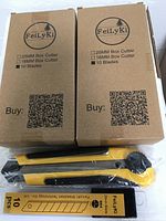 Two unopened brown cardboard boxes labeled FeiLyKi containing yellow and black box cutters and blade refills. Each box lists 18mm box cutter and 10 blades.
