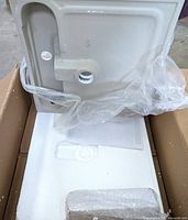 Bottom side of the white ceramic vessel sink showing drain hole and mounting points, partially wrapped in plastic inside packaging box.
