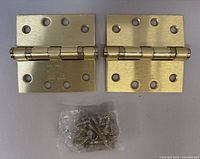 Two satin brass square door hinges with 8 holes each, placed side by side on a light background with screws in a sealed plastic bag in front.
