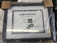 Three black certificate frames stacked, each contains a certificate placeholder showing the size and decorative mat border, wrapped in protective plastic and cardboard corner guards.