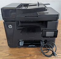 Front view of the black HP Laser Jet Pro MFP M225dw wireless monochrome printer, showing the paper output tray, HP branding, and 3-inch touchscreen display with attached power cable.