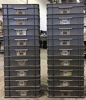 Two stacks of grey plastic crates, approximately 10 in each stack, showing barcode labels on sides and overall condition.