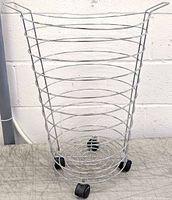 Front view of the stainless steel wired laundry basket showing round wire loops and caster wheels on a concrete floor against a white wall.