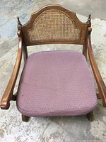 Front view of vintage wooden chair with cane back and mauve upholstered seat showing the carved armrests and distinctive cane panel in the backrest.