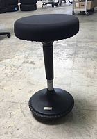 Side view of black wobble stool showing round fabric seat, central adjustable support column, and wide round base with rubber edge.