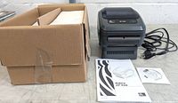 Zebra ZP450 thermal label printer with power cord, box for labels, user manual and software CD arranged flat on a surface.