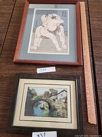 Two framed prints displayed side by side on a wooden surface with a measurement ruler. The top print depicts a sumo wrestling scene; the bottom print shows a canal with a bridge and boat.