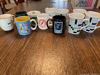 Front view of all nine coffee mugs lined up on wooden table showing variety of designs including crossword grids, colorful prints and solid colors.