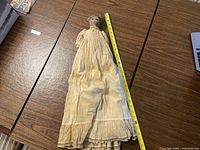 Full body view of antique fabric doll on wooden table. Doll wears a long white dress, measuring approximately 24 inches tall, showing overall wear and discoloration.