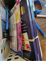 Photo showing packs of colored pencils, Sharpie markers, and transparent case with colored markers along with wooden boxes in the background.