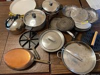 Large assortment of cooking pans, pots, splatter screens, steamers and lids arranged on wooden tables, showing yellow Dansk Kobenstyle paella pan, copper pan, cast iron cornbread pan, stainless steel pots with lids, metal steamers and splatter screens.