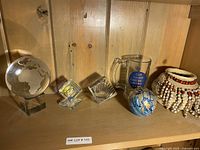 Overall view of assorted decorative items on wooden shelf including glass globe, glass cube paperweight, small glass shell, glass tankard, blue ceramic ball, and beaded tribal-style necklace.