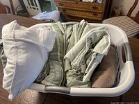 Laundry basket containing folded flannel sheets, linens including a plaid green pillowcase and solid colored sheets, and a brown pillow