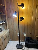 Full view of the black 3 light floor lamp standing against a wood paneled wall, showing the lamp lit on one of the heads and the round base and cord on the floor.