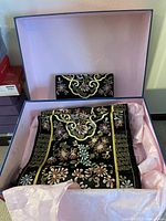 Both clutch and scarf placed in pink-lined box, showing floral embroidery and gold detailing on black fabric.