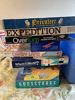 Stack of board games including Privateer, Expedition, Overturn, Candy Land, Where in the World?, Guesstures, and Trivial Pursuit, visible box edges, titles and designs
